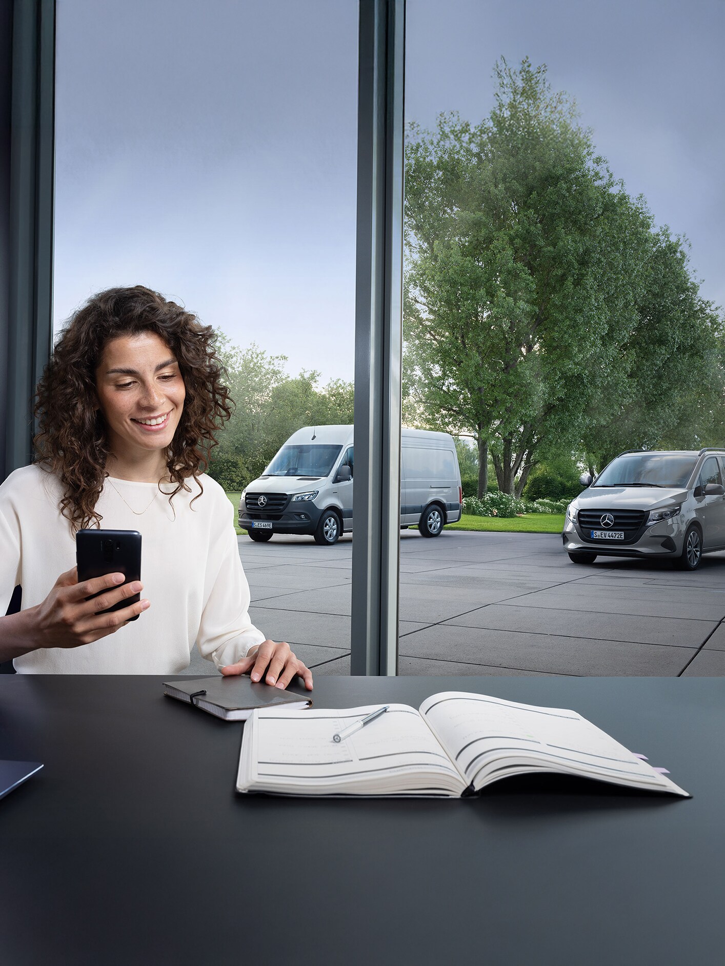 Pencere önünde akıllı telefonuyla bir kadın ve dışarıda üç Mercedes-Benz hafif ticari araç.