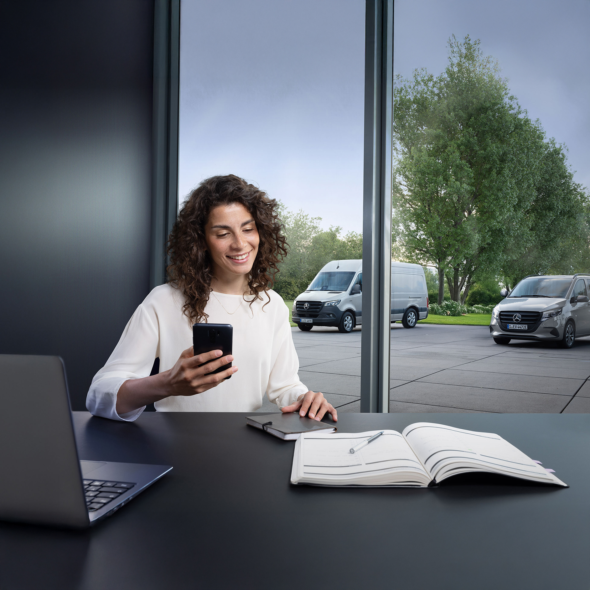 Mercedes-Benz | Konfor | Çevrimiçi randevu rezervasyonu Pencere önünde akıllı telefonuyla bir kadın ve dışarıda üç Mercedes-Benz hafif ticari araç.