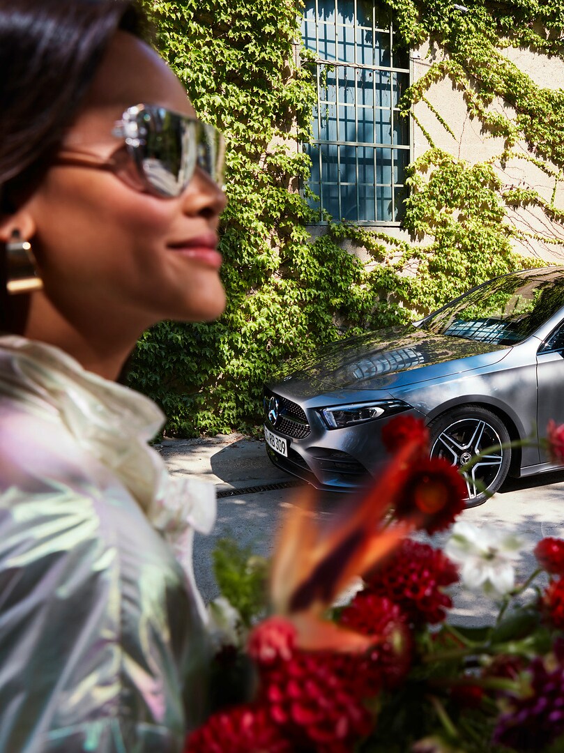 Rahatlamış bir kadın elinde çiçeklerle bakımı yapılmış Mercedes'inin yanına gidiyor.