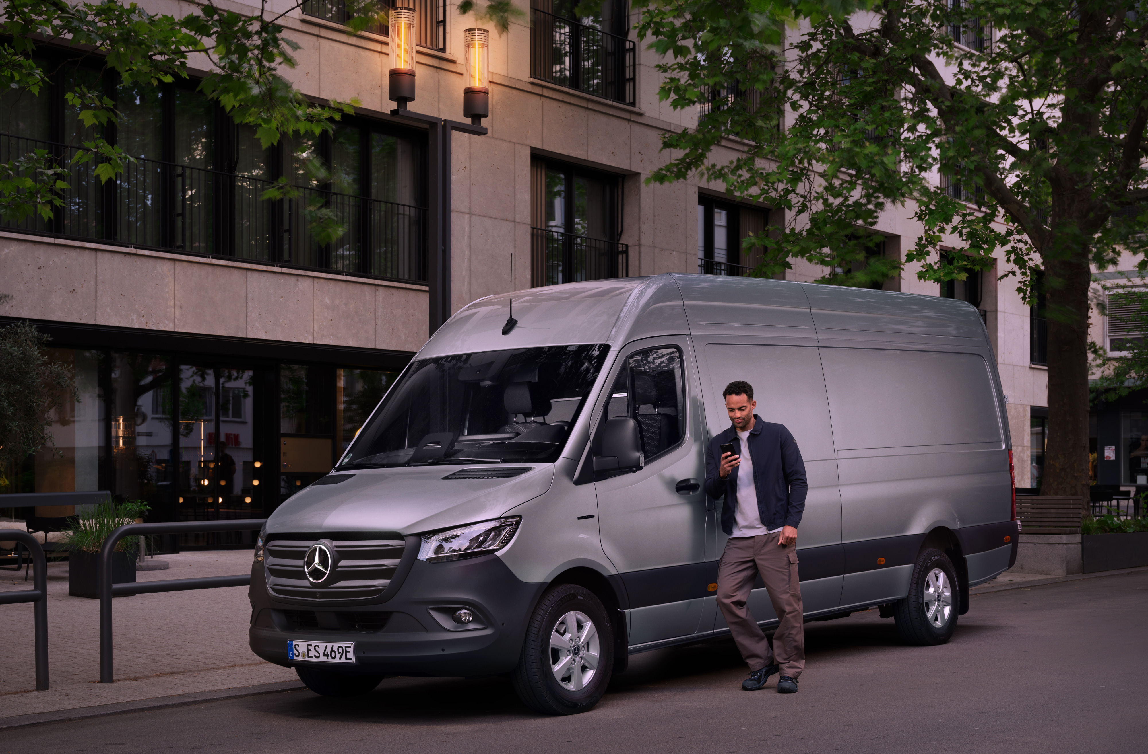 Mercedes-Benz | Hafif Ticari Araç Bakımı | Müşteri İletişim Merkezi Bir erkek gümüş Mercedes-Benz Sprinter hafif ticari araca yaslanıyor ve akıllı telefonunda Mercedes Benz Müşteri Hizmetlerinden hızlı desteği kullanıyor.