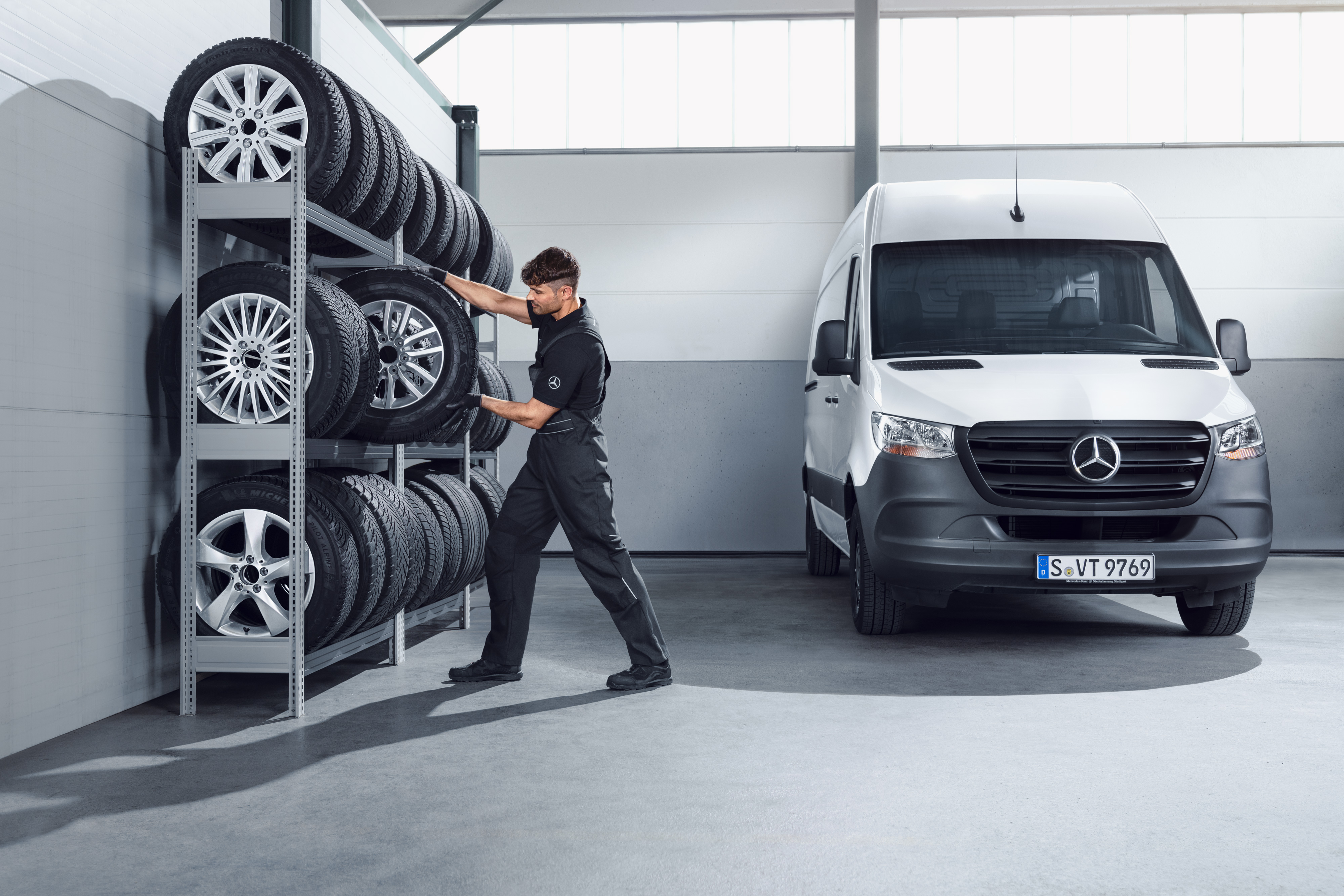 Mercedes-Benz | Hafif Ticari Araçlar Orijinal Parça Kalitesi Beyaz bir Sprinter için servis rafından Mercedes-Benz komple kış lastikleri seçen bir mekanik teknisyeni.