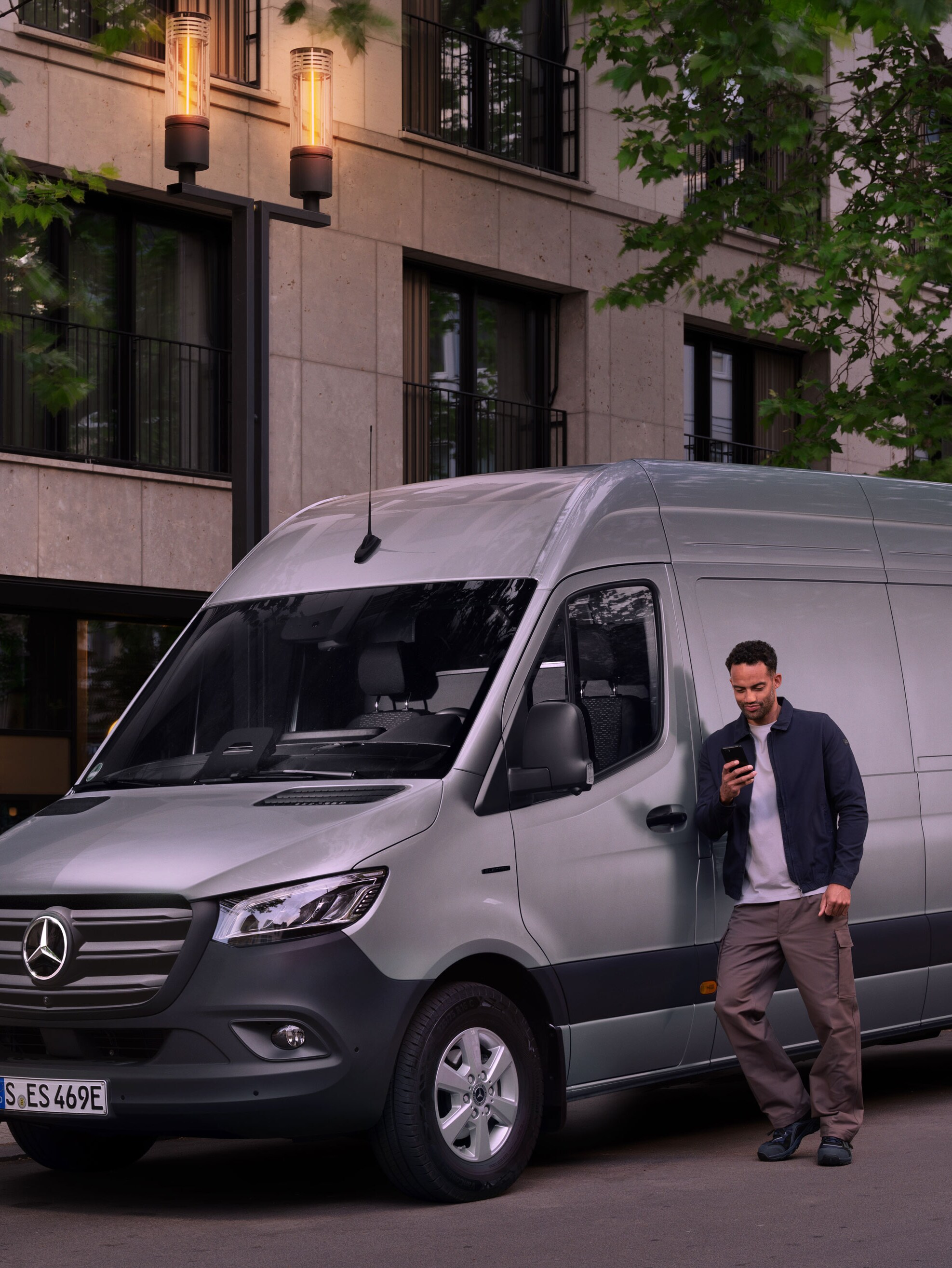 Bir adam gümüş bir Mercedes-Benz Sprinter'a yaslanıyor ve mobil telefonundan hasar raporunu tamamlıyor.